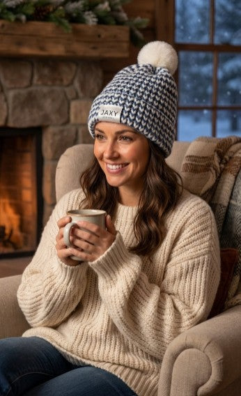 Woman wearing a jaxy beanie and sweater, holding a mug in front of a fireplace.
