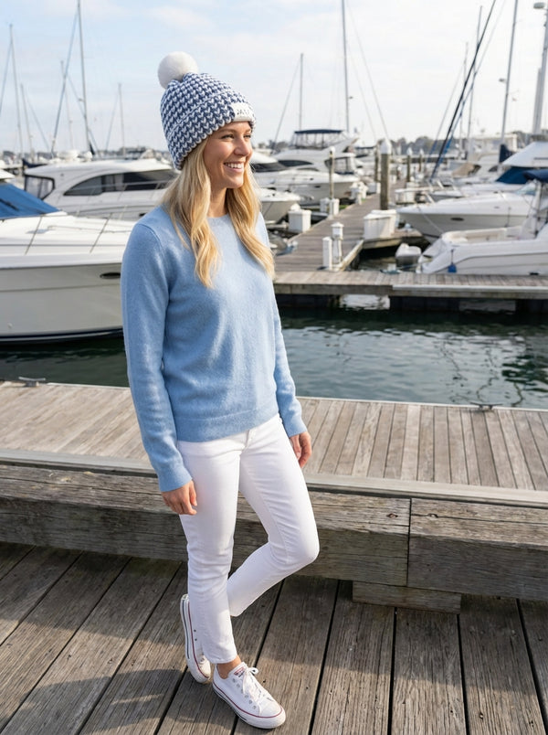 Woman in light blue sweater wearing sky jaxy beanie and white pants standing on a dock with boats in the background
